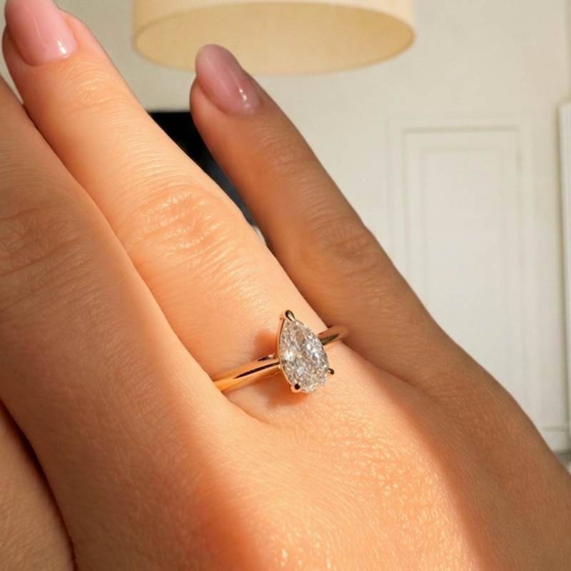 A gold ring with a pear-cut clear gemstone worn on a hand with pink manicured nails, set against a softly blurred indoor background with a lamp.